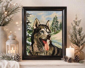 May include: A framed needlepoint tapestry depicting a husky dog with blue eyes and a red tongue hanging out. The dog is set against a winter landscape featuring snow-covered pine trees and a light blue sky, all enclosed in a black frame.