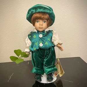Puede incluir: Una muñeca de porcelana vestida con un atuendo verde con patrones de trébol. La muñeca lleva un sombrero verde y sostiene un trébol.