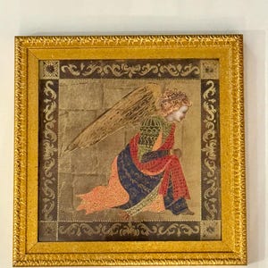 May include: A framed artwork featuring a kneeling angel with a large wing. The angel wears a colorful robe in red, blue, and green. The artwork is set against a gold background and framed in a gold ornate frame.