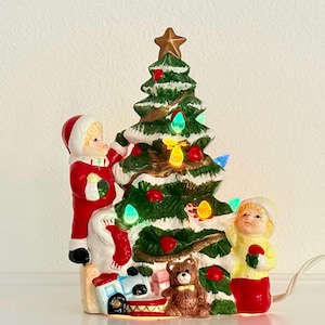 May include: A ceramic Christmas tree with a gold star topper, decorated with colorful lights and red ornaments. Two children in festive attire stand beside the tree, with a teddy bear and toy train at the base. The tree is lit with a power cord.