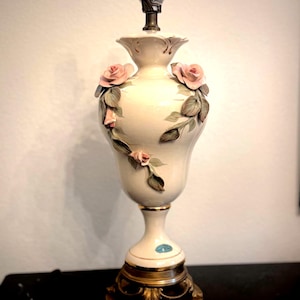 May include: A cream-colored ceramic table lamp base with a bulb socket at the top. The body features three-dimensional pink rose blossoms and green leaves applied to the surface. It rests on a circular, ornate gold-toned metal pedestal base.