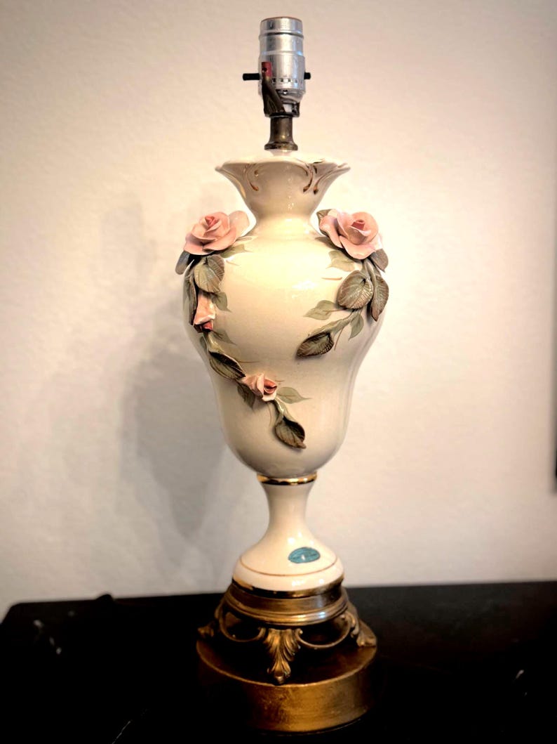 May include: A cream-colored ceramic table lamp base with a bulb socket at the top. The body features three-dimensional pink rose blossoms and green leaves applied to the surface. It rests on a circular, ornate gold-toned metal pedestal base.