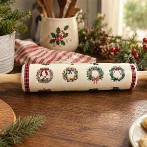 May include: A cylindrical rolling pin with a cream-colored barrel featuring a repeating pattern of various holiday wreaths and red plaid trim at both ends. The rolling pin has smooth, light-toned wooden handles on each side for gripping.