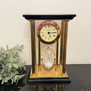 Pode incluir: Um suporte de relógio e ampulheta dourado com uma base preta. O mostrador do relógio tem algarismos romanos e o texto "Grand Palais" no centro. A ampulheta está cheia de areia.