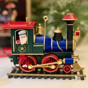 May include: A miniature Christmas train ornament. The train is painted in green, blue, red, and gold. Santa Claus is in the driver's seat. The train is on a black track. The text "SANTA'S SPECIAL" is on the side of the train.