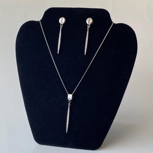 May include: A silver-toned jewelry set displayed on a black velvet stand. The set includes a necklace with a rectangular pendant and a dangling, elongated design, and matching earrings. The earrings and pendant feature a line of small, sparkling stones.