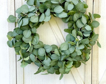May include: A green eucalyptus wreath with small white flowers. The wreath is hanging on a white wooden door.