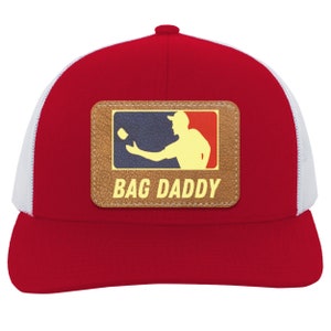 May include: Red and white trucker hat with a brown leather patch featuring a silhouette of a person throwing a beanbag with the text "BAG DADDY" underneath.