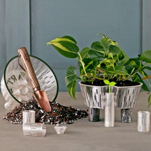 May include: A set of silver-coloured, perforated metal planters with a small green plant in one of the planters. A small trowel, a pile of brown potting soil, and additional planters are also shown.