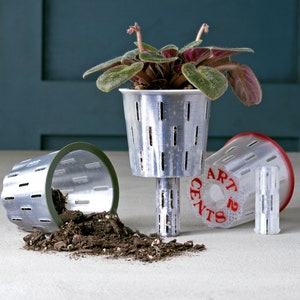 Peut inclure: Trois pots de fleurs en métal argenté avec des trous de drainage. Le pot du centre a une plante verte avec des feuilles violettes. Le pot de droite porte l'inscription "ART 2 CENTS". Le pot de gauche est renversé et de la terre se répand.
