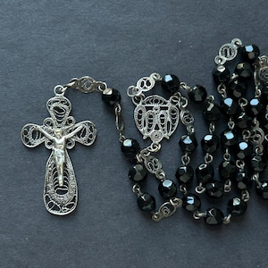 May include: A silver filigree crucifix with a detailed depiction of Jesus Christ, connected to a strand of faceted black beads. The rosary is laid out against a dark background, showcasing its intricate design.