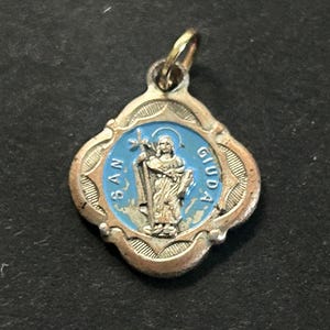May include: A metallic pendant featuring a central light blue enamel inlay with a raised silver-toned figure of a person holding a staff. The text SAN GIUDA is inscribed on the blue background. The pendant has a decorative scalloped border.