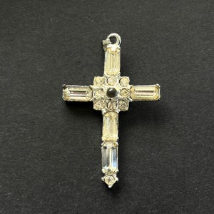 May include: A silver-toned metal cross pendant featuring clear baguette-cut and round-cut rhinestones. The center of the cross is adorned with a cluster of round stones surrounding a single dark circular accent, with a small loop at the top.