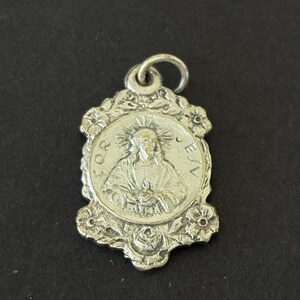 May include: A silver-toned metal pendant featuring a central embossed figure of Jesus with a halo, surrounded by a floral border. The text COR JESU is inscribed on the sides of the central circular design. A small metal jump ring is attached to the top.