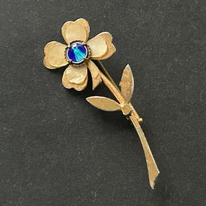 May include: A gold-toned flower brooch with a blue and teal center stone. The brooch is in the shape of a flower with four petals and a stem with two leaves. The brooch is likely made of metal.