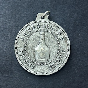 May include: Silver-toned metal keychain featuring a round pendant with the words "Benedictine Fecamp France" and a stylized image of a bottle of liqueur.