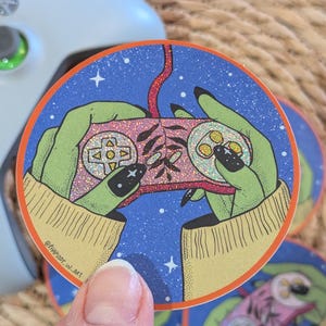 Könnte beinhalten: Ein runder Aufkleber mit blauem Hintergrund und einem rosa-roten Videospiel-Controller, der von zwei grünen Händen mit schwarzen Fingernägeln gehalten wird. Der Aufkleber hat eine glitzernde Textur und Sterne im Hintergrund. Der Text "@HARPSODY_IN_ART" befindet sich am unteren Rand des Aufklebers.