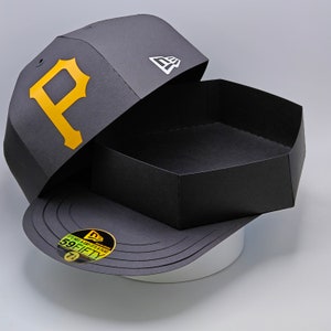 May include: A black baseball cap with a gold "P" logo and a white New Era logo. The cap is open to reveal a black box with a white "59FIFTY" sticker.