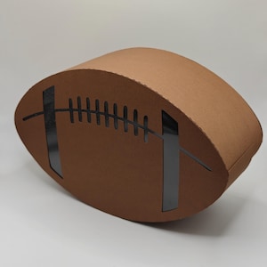 May include: A brown, football-shaped cardboard box featuring a cutout design that mimics the laces and stripes of an American football. The box has a smooth, matte finish and a curved, three-dimensional structure with visible corrugated edges.