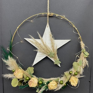 Könnte beinhalten: Ein goldener Metallreifenkranz mit getrockneten Blumen, Grün und Pampasgras. Ein weißer Holzstern mit einem glitzernden grünen Stern-Akzent ist am Kranz befestigt.