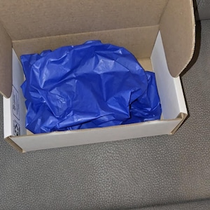 May include: Open cardboard box with a white interior, filled with crumpled blue tissue paper. The box is on a gray surface.