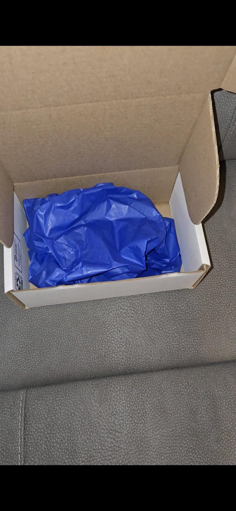 May include: Open cardboard box with a white interior, filled with crumpled blue tissue paper. The box is on a gray surface.