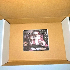 May include: A square sticker inside a cardboard box. The sticker features Santa Claus in his traditional red suit, seated in a chair, giving the middle finger. He holds a glass of milk and a plate of cookies.