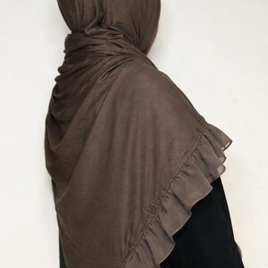 May include: A brown hijab with a ruffled edge. The head covering is draped over the head and shoulders, with the ruffled trim adding a decorative element. The hijab is made of a soft, flowing fabric.