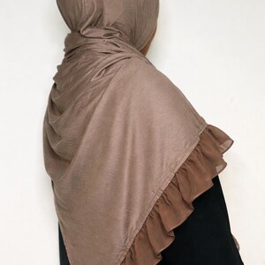 May include: A brown hijab with a ruffled trim. The head covering is made of a soft fabric and drapes over the shoulders. The hijab is a solid color and is worn over a black garment.