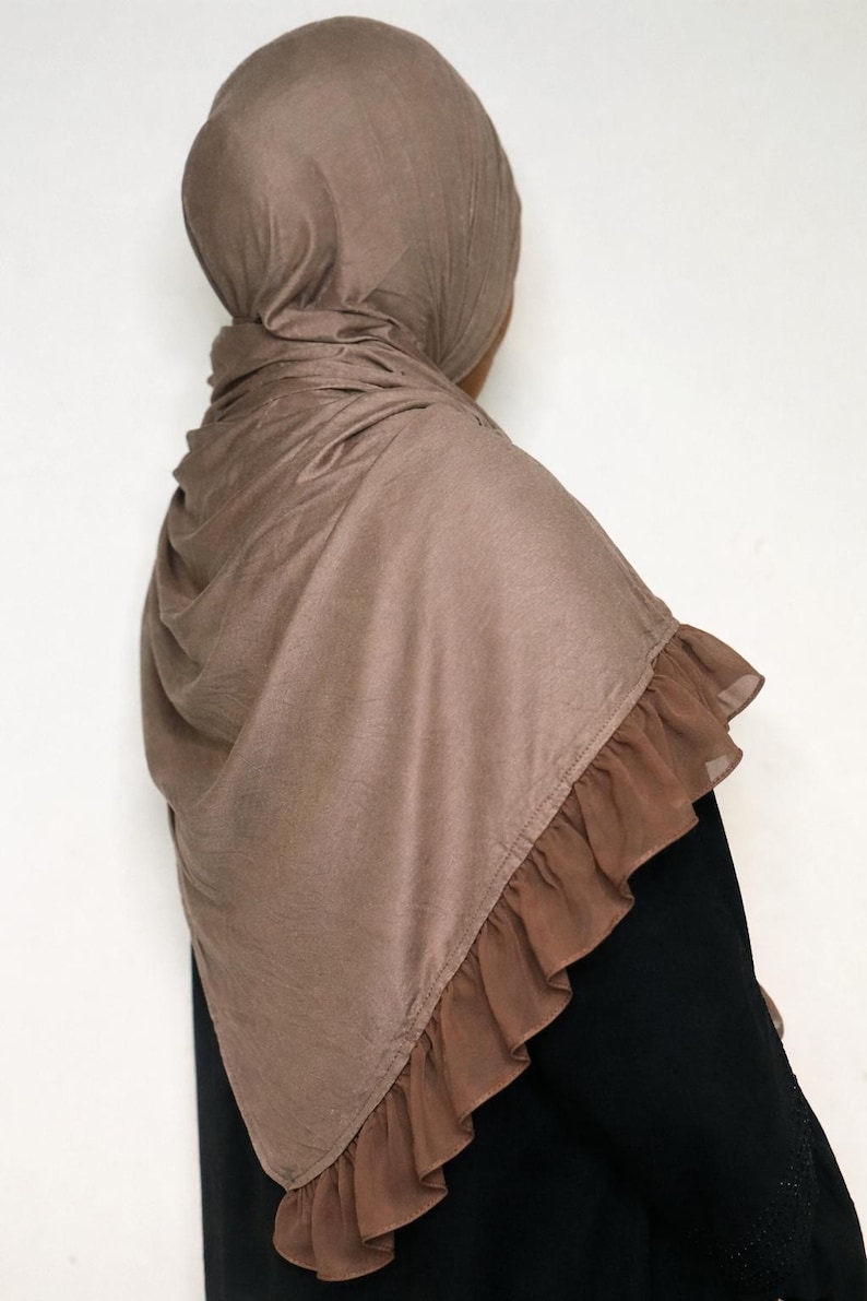 May include: A brown hijab with a ruffled trim. The head covering is made of a soft fabric and drapes over the shoulders. The hijab is a solid color and is worn over a black garment.