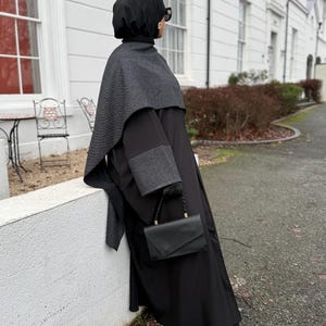 Puede incluir: Un conjunto negro que presenta un vestido largo, una capa estampada y un tocado a juego. El atuendo incluye un pequeño bolso de mano negro con un patrón geométrico. La persona lleva guantes y gafas de sol negros. El fondo es un edificio blanco.