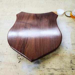 May include: A shield-shaped wooden plaque with a polished, dark brown finish. The plaque has a smooth surface and a curved top edge. The wood grain is visible, adding texture to the piece. The plaque is likely for display or engraving.