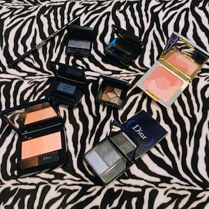 May include: An assortment of makeup products, including eyeshadow palettes and blush compacts, are displayed on a zebra-print surface. The products are in various shades of pink, brown, and gray, with some featuring mirrors and applicators. A black eyeliner pencil is also present.