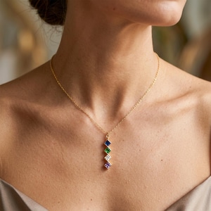 Puede incluir: Un collar dorado con una cadena fina. El colgante presenta cinco piedras cuadradas en azul, verde, blanco, morado y rosa. El collar se lleva alrededor del cuello.