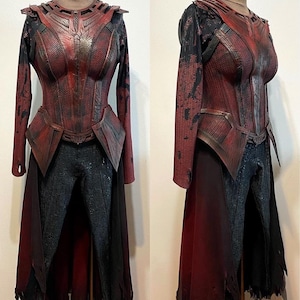 Pode incluir: Um traje de duas peças exibido em um manequim. A parte superior é um corpete vermelho e preto com mangas compridas, e a parte inferior são calças cinza escuro. O traje tem uma aparência desgastada e texturizada, com uma capa presa à cintura.