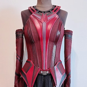 Op de afbeelding: Een rood en zwart kostuum met een geometrisch ontwerp. Het kostuum heeft een hoge halslijn en lange mouwen. De stof is textuur en heeft een metallic glans.