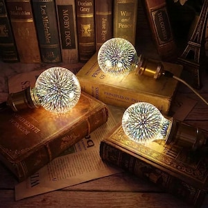 May include: Three spherical LED light bulbs with a multi-coloured 3D firework effect glass finish. Each bulb features a metallic brass-toned base and is positioned resting on top of vintage hardcover books with visible text and spines.
