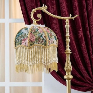 May include: A floor lamp with a gold-toned metal stand and a curved arm. The lampshade features a multi-colored floral tapestry fabric with a scalloped edge, gold braided trim, and a long fringe of hanging white beads and gold threads.