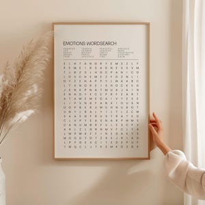 May include: A framed Emotions Wordsearch art print. The print features a grid of letters with words related to emotions listed above. The frame is a light brown color, and the print is displayed on a white wall.