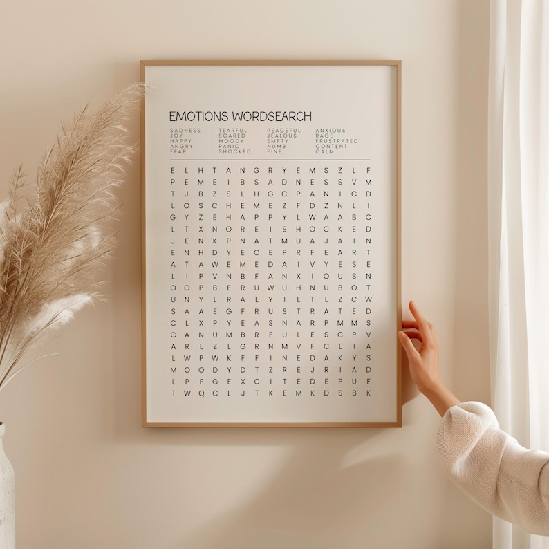 May include: A framed Emotions Wordsearch art print. The print features a grid of letters with words related to emotions listed above. The frame is a light brown color, and the print is displayed on a white wall.