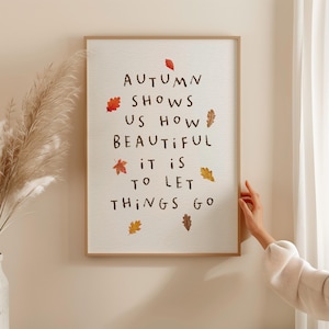 Puede incluir: Una impresión artística enmarcada con el texto "AUTUMN SHOWS US HOW BEAUTIFUL IT IS TO LET THINGS GO". La impresión presenta hojas de otoño en tonos rojos, naranjas y marrones. El marco es de color marrón claro.
