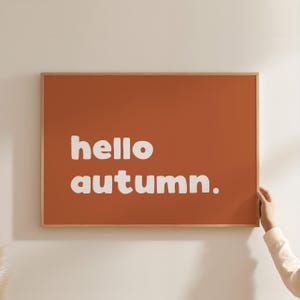Könnte beinhalten: Ein gerahmter rechteckiger Kunstdruck mit einem orangefarbenen Hintergrund. Die Worte "hello autumn." sind in weißen, fetten, kleinen Buchstaben geschrieben. Der Rahmen ist hellbraun. Der Druck wird von einer Person gehalten.