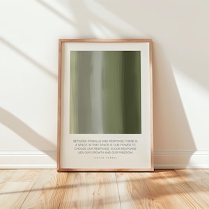 May include: A framed print featuring an abstract design of vertical brushstrokes in shades of green and gray. The print includes a quote by Viktor Frankl: "Between stimulus and response..." The frame is a light brown color.