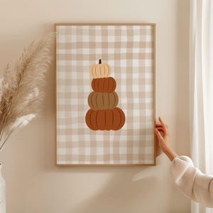 Op de afbeelding: Een ingelijste print met een stapel pompoenen in tinten oranje en bruin. De pompoenen zijn geplaatst tegen een beige en witte geruite achtergrond. Het kunstwerk wordt gepresenteerd in een houten lijst.
