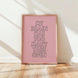 May include: A framed print with the text "MY BRAIN HAS TOO MANY TABS OPEN" in black letters on a pink background. The frame is a light brown color, and the print is displayed on a wooden floor.