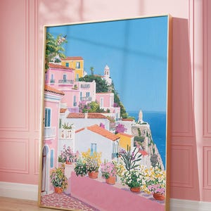 Op de afbeelding: Een ingelijste kunstdruk met een kustdorpsscène. Het kunstwerk toont roze gebouwen met oranje daken, weelderige bloemen en een heldere blauwe lucht. De print wordt tentoongesteld tegen een roze muur.