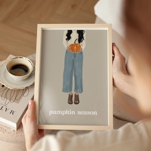 May include: A framed art print featuring a person holding a pumpkin. The person is wearing a cream-colored sweater, blue pants, and brown boots. The words "pumpkin season" are printed in white at the bottom of the print.