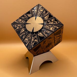 May include: A black and gold cube puzzle with intricate designs on each side. The cube is on a silver stand.