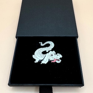 Puede incluir: Un pin de esmalte de dragón blanco y plateado con una lengua rosa, presentado en una caja de regalo negra. El pin presenta un diseño de dragón estilizado con detalles intrincados. La caja tiene una cinta negra.