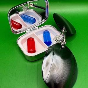 May include: A silver pill case with a mirror, containing red and blue capsules. A dark green, oval-shaped accessory hangs from the case. The background is a solid green.